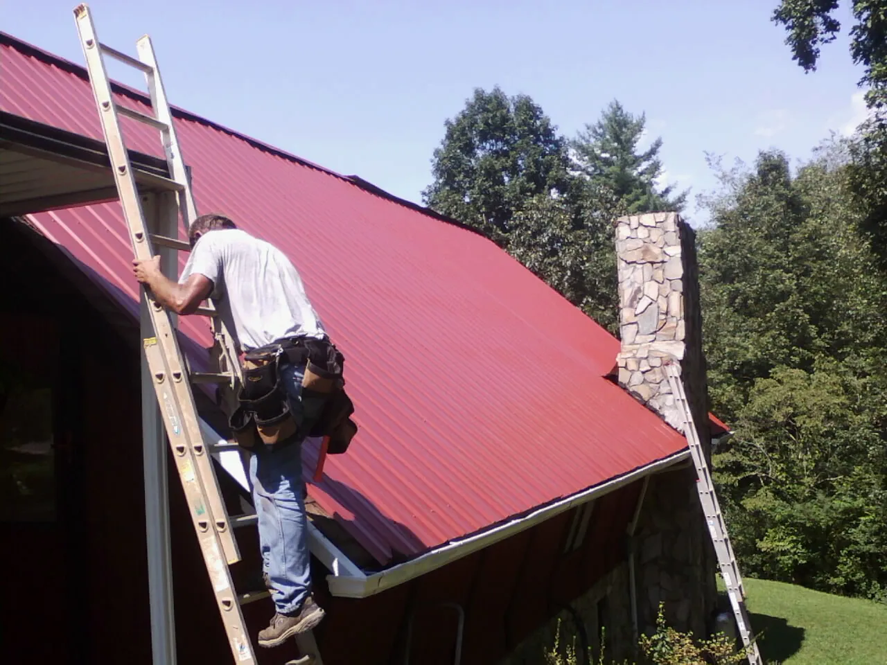 Roofer on a ladder beside a red metal roof and stone chimney — Soffit & Fascia Repair by Roofing Brothers in Hellertown